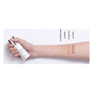 Eine Hand hält ein Fläschchen Foundation neben farbigen Hauttönen auf dem Unterarm, die verschiedene Nuancen zeigen.