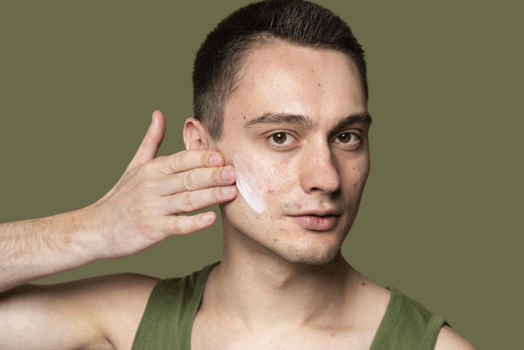 Ein Mann trägt Gesichtscreme auf seine linke Wange auf, Hintergrund ist ein einfarbiger, olivgrüner Farbton.