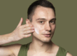 Ein Mann trägt Gesichtscreme auf seine linke Wange auf, Hintergrund ist ein einfarbiger, olivgrüner Farbton.