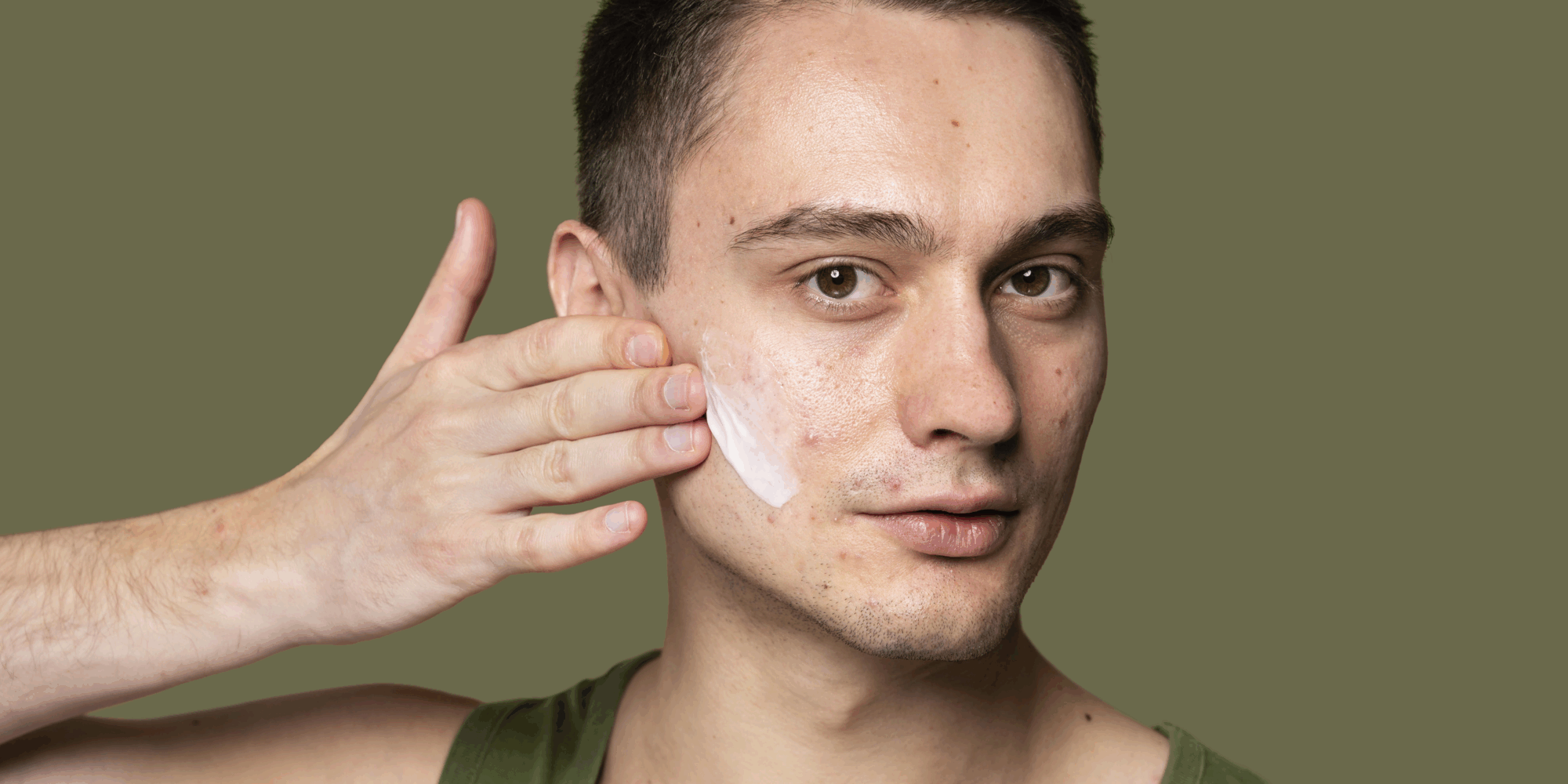 Ein Mann trägt Gesichtscreme auf seine linke Wange auf, Hintergrund ist ein einfarbiger, olivgrüner Farbton.