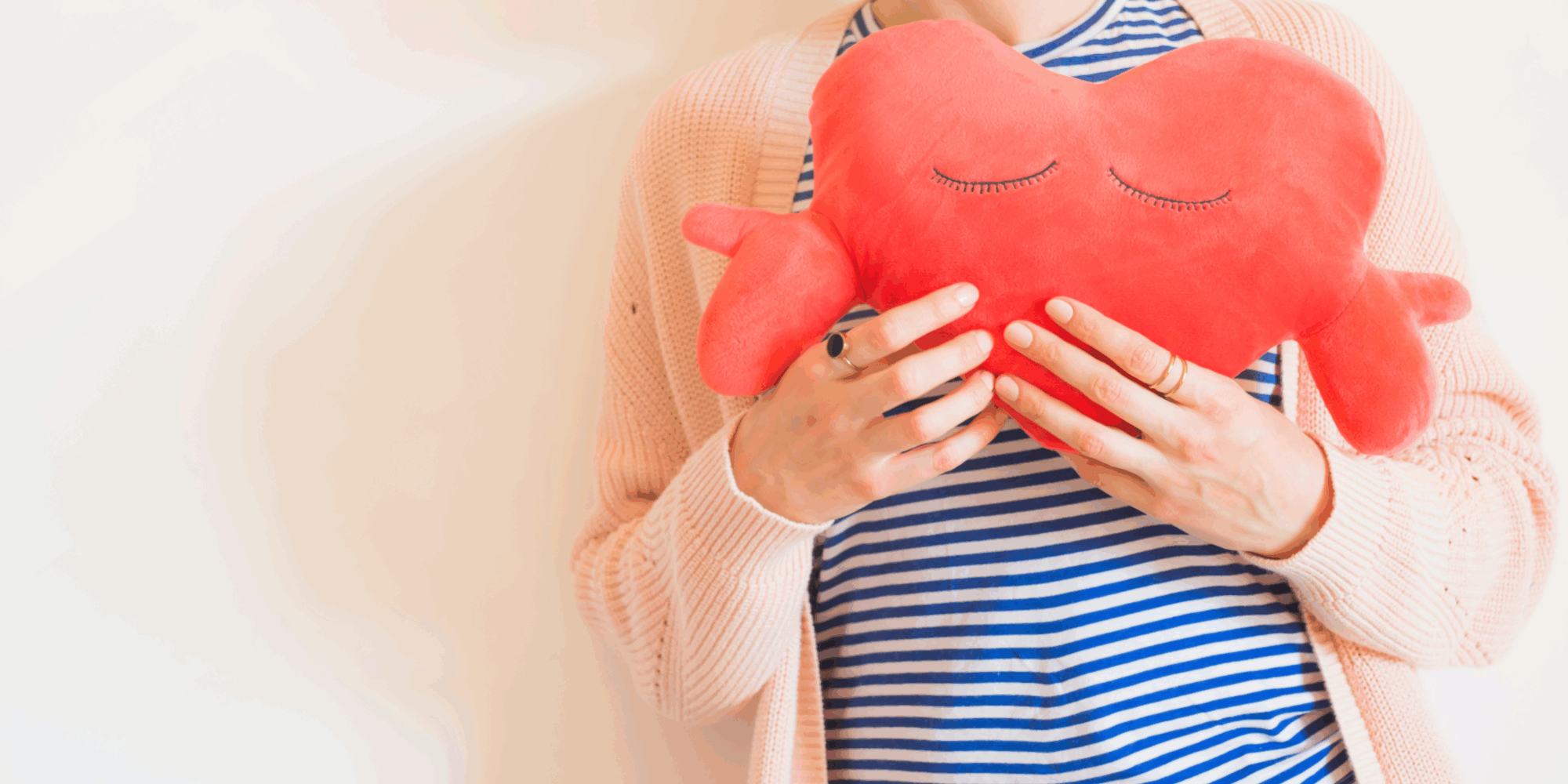 Herzinfarkt_Frauen.png Eine Person hält ein rotes herzförmiges Kissen mit geschlossenen Augen, gekleidet in gestreiftes Oberteil und eine helle Strickjacke.