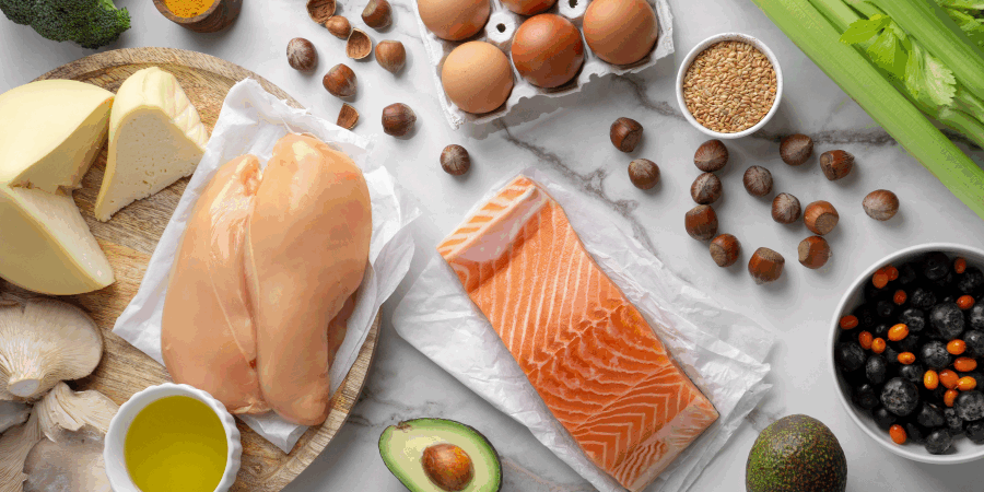 Verschiedene Lebensmittel wie Hähnchenbrust, Lachs, Käse, Eier, Avocado und Gemüse auf einem Tisch.