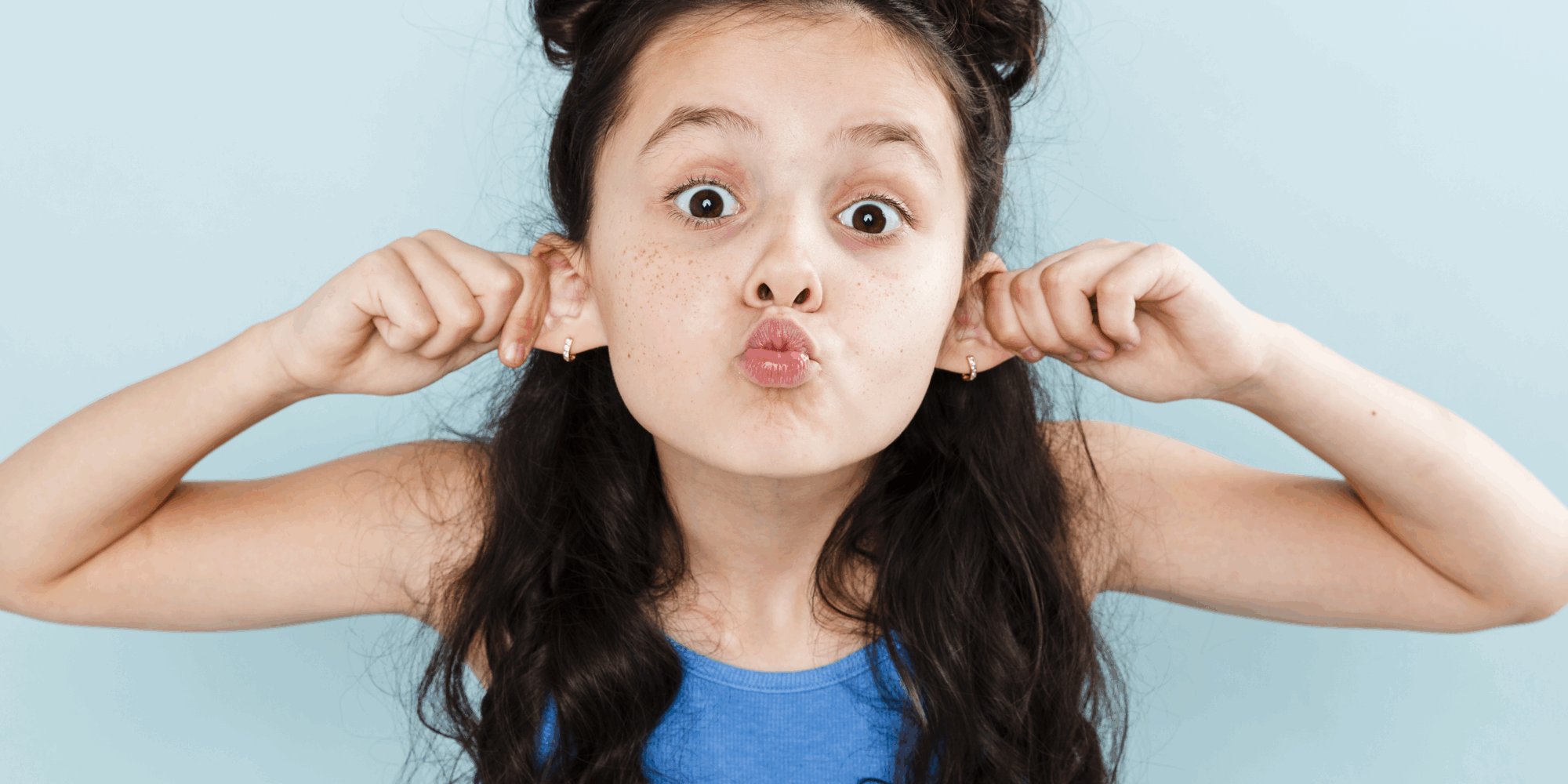 Ein Mädchen mit langen, lockigen Haaren zieht an ihren Ohren und macht ein lustiges Gesicht vor einer hellblauen Wand.