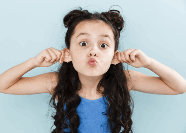 Ein Mädchen mit langen, lockigen Haaren zieht an ihren Ohren und macht ein lustiges Gesicht vor einer hellblauen Wand.