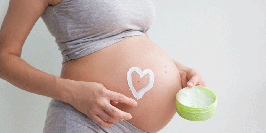 Eine schwangere Person trägt eine Crememaskierung in Herzform auf ihren Bauch auf, während sie eine grüne Dose hält.