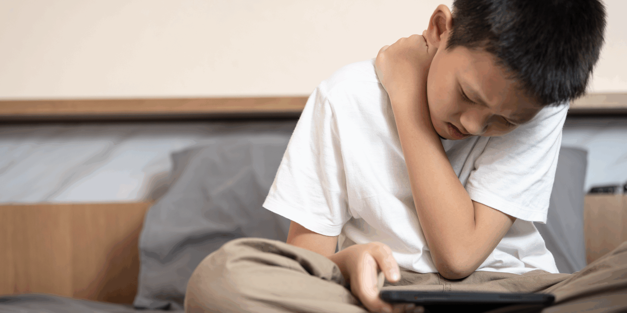 Ein Junge sitzt auf einem Bett, hält ein Tablet und fasst sich schmerzverzerrt an den Hals.