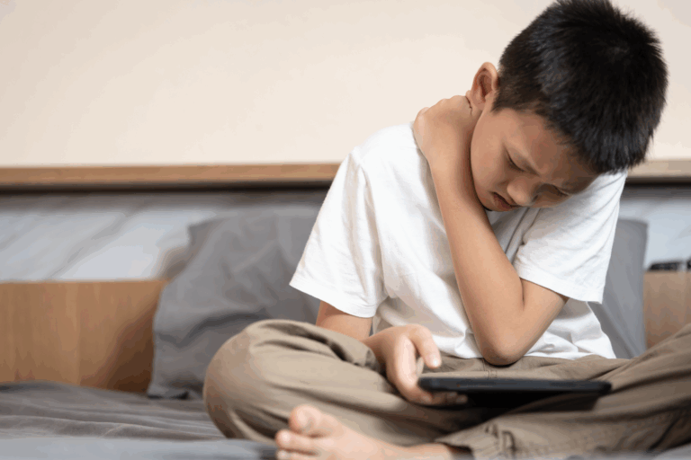 Ein Junge sitzt auf einem Bett, hält ein Tablet und fasst sich schmerzverzerrt an den Hals.