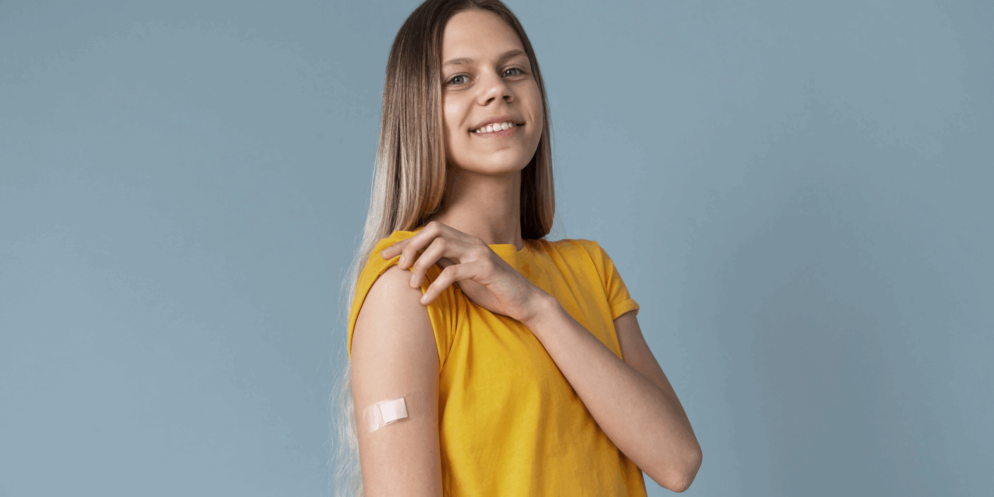 Eine lächelnde junge Frau mit langen Haaren trägt ein gelbes T-Shirt und zeigt einen Verband am Oberarm.