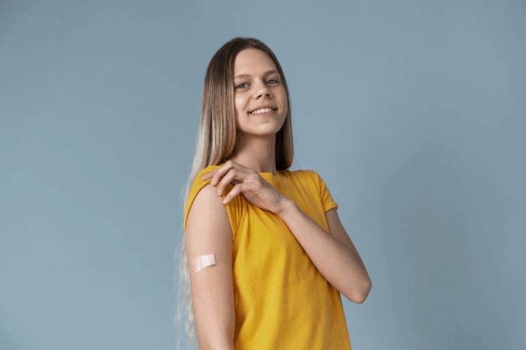 Eine lächelnde junge Frau mit langen Haaren trägt ein gelbes T-Shirt und zeigt einen Verband am Oberarm.