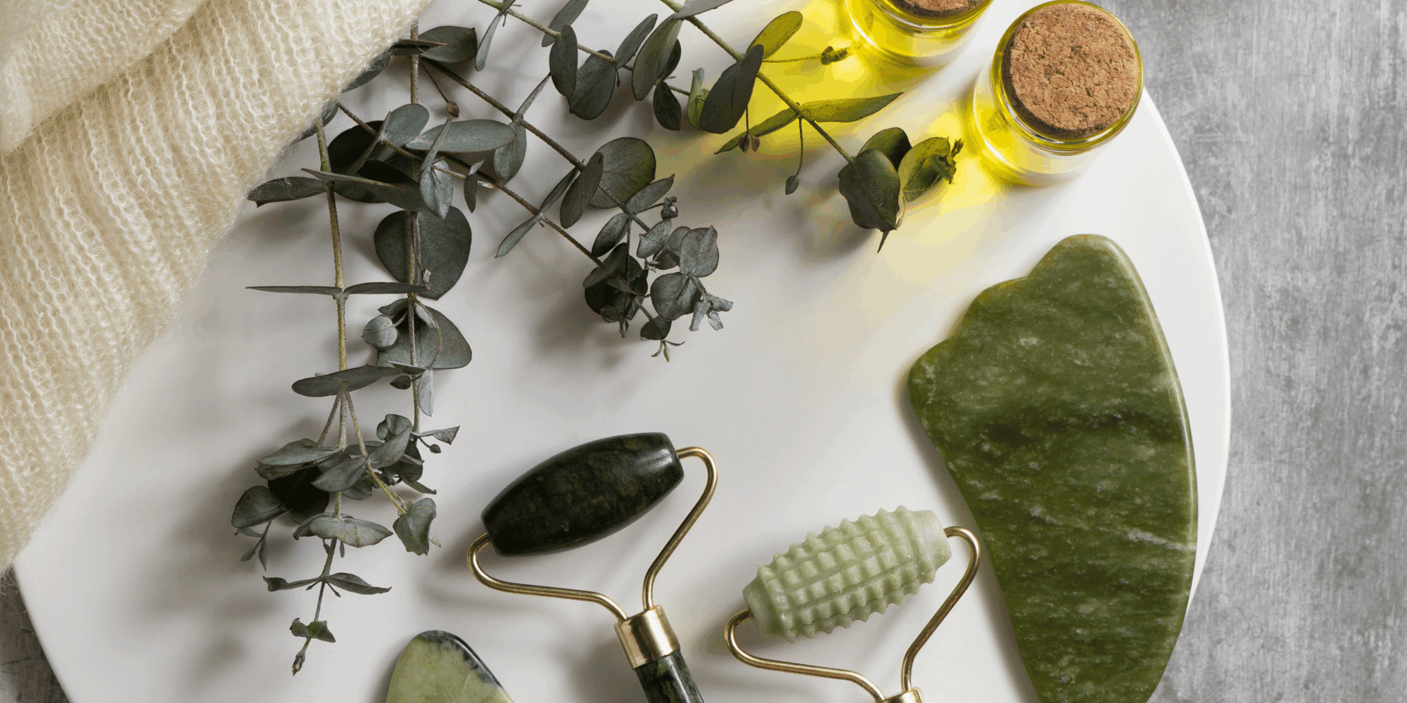 nachhaltige-kosmetik.png Ein weißer Teller mit drei Jade-Rollern, einem gua sha Stein, zwei Fläschchen ätherischem Öl und Eukalyptuszweigen.