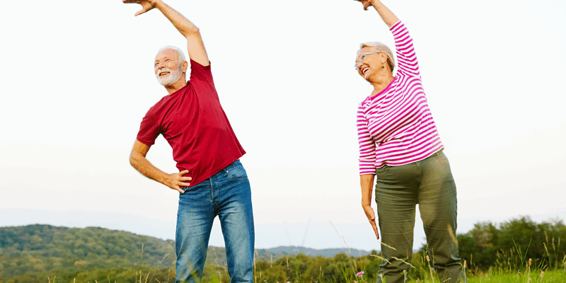 Ein älteres Paar führt Dehnübungen im Freien auf einer Wiese durch, umgeben von Landschaft und Hügeln.