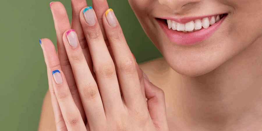 Nahaufnahme einer lächelnden Person mit bunten Nagelspitzen und sichtbaren, gepflegten Händen vor grünem Hintergrund.
