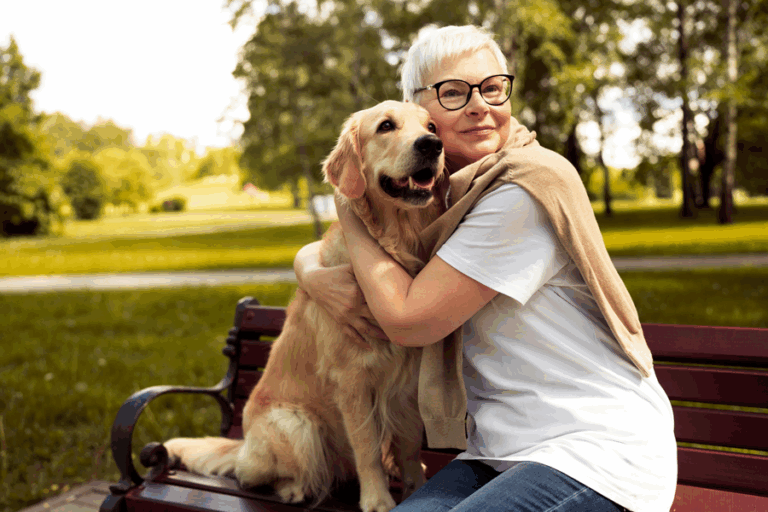 Eine Frau mit kurzen, blonden Haaren und Brille umarmt einen golden retriever auf einer Bank im Park.