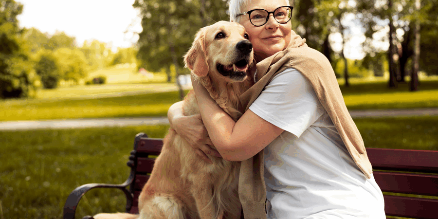 Hunde-als-Seelentroester.png Eine Frau mit kurzen, blonden Haaren und Brille umarmt einen golden retriever auf einer Bank im Park.