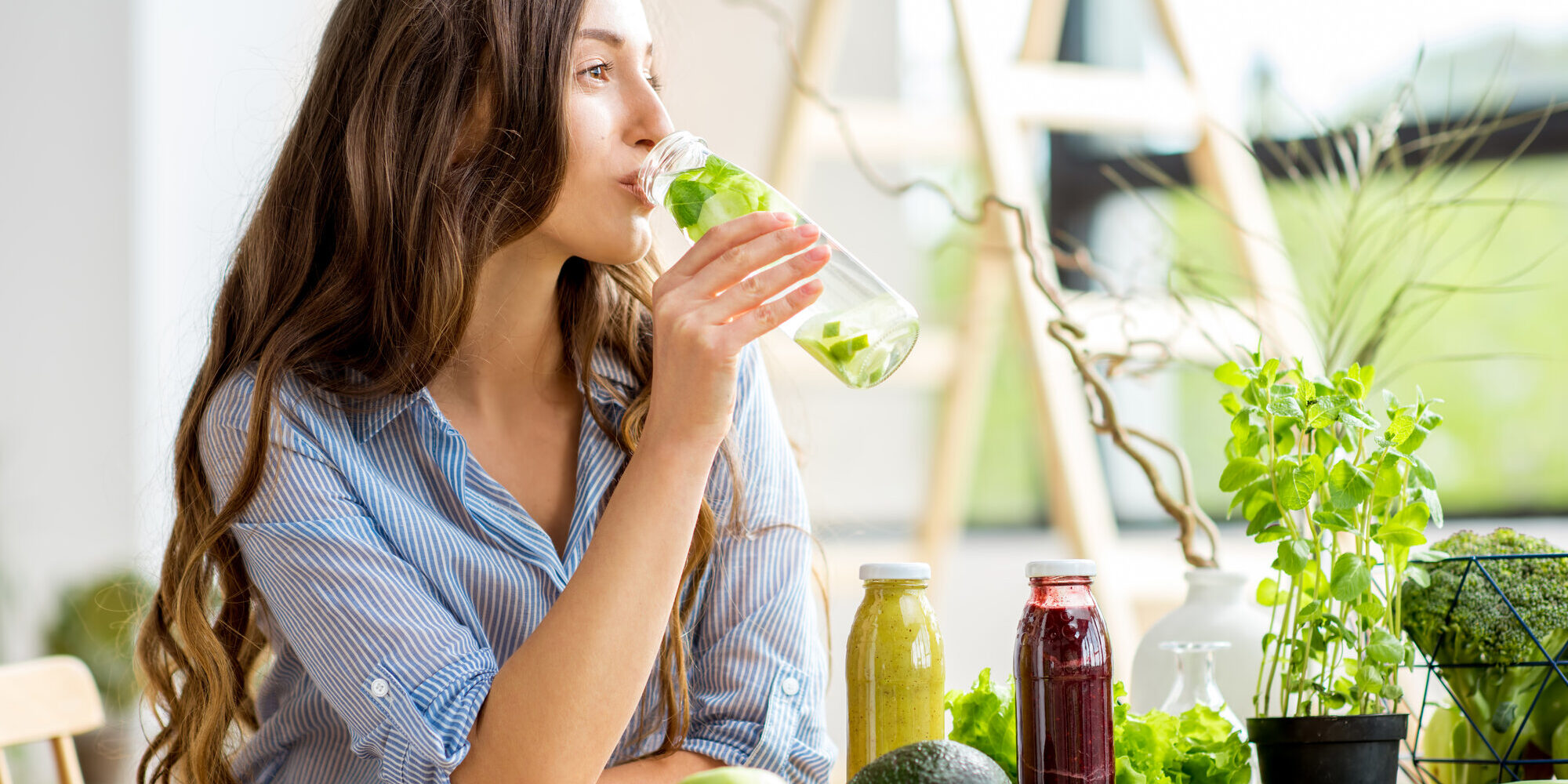 Saeure-Basen-Haushalt-Balance.jpg Eine Frau trinkt aus einer Glasflasche mit frischer Minze, umgeben von gesunden Lebensmitteln und Pflanzen.