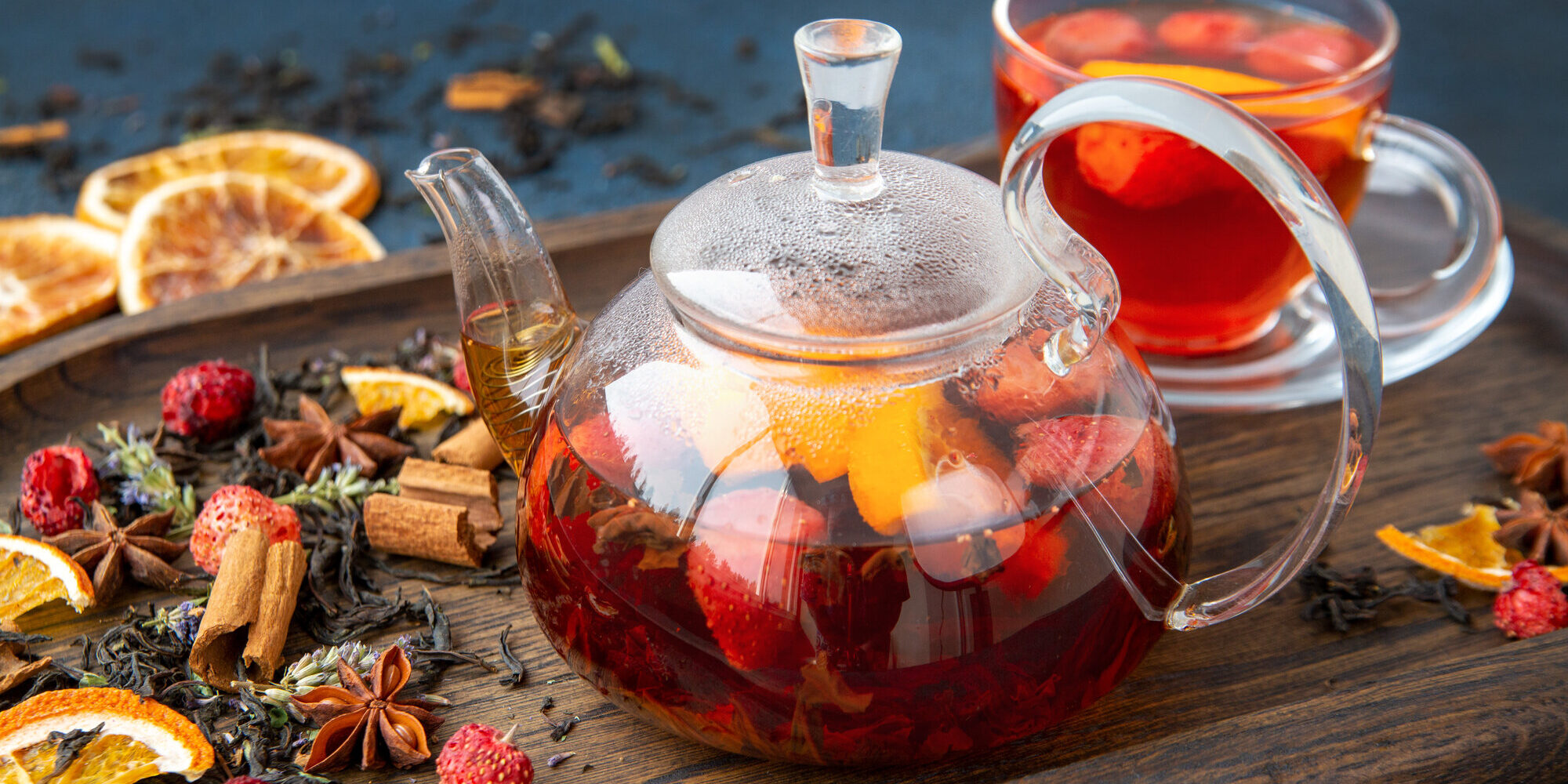 Teekanne aus Glas mit fruchtigem Tee, umgeben von Trockenfrüchten und Gewürzen auf einem Holzbrett.