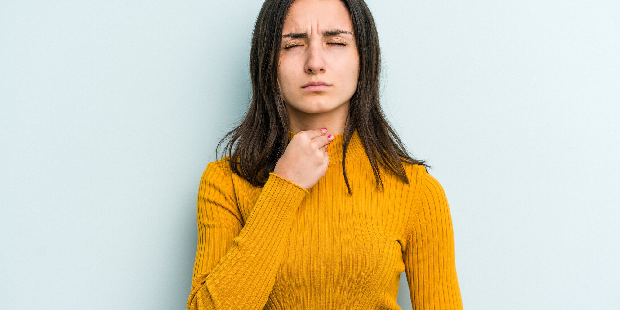 Stimme_versagt.jpg Frau in gelbem Pullover, die nachdenklich auf ihre Kehle zeigt, vor einer hellblauen Wand.