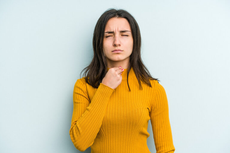 Frau in gelbem Pullover, die nachdenklich auf ihre Kehle zeigt, vor einer hellblauen Wand.
