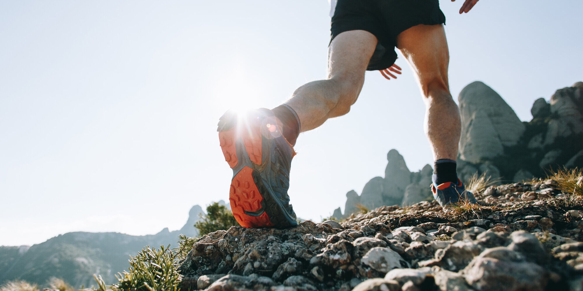 Läufer in sportlichen Schuhen auf einem kies- und steinigen Weg mit Berglandschaft im Hintergrund
