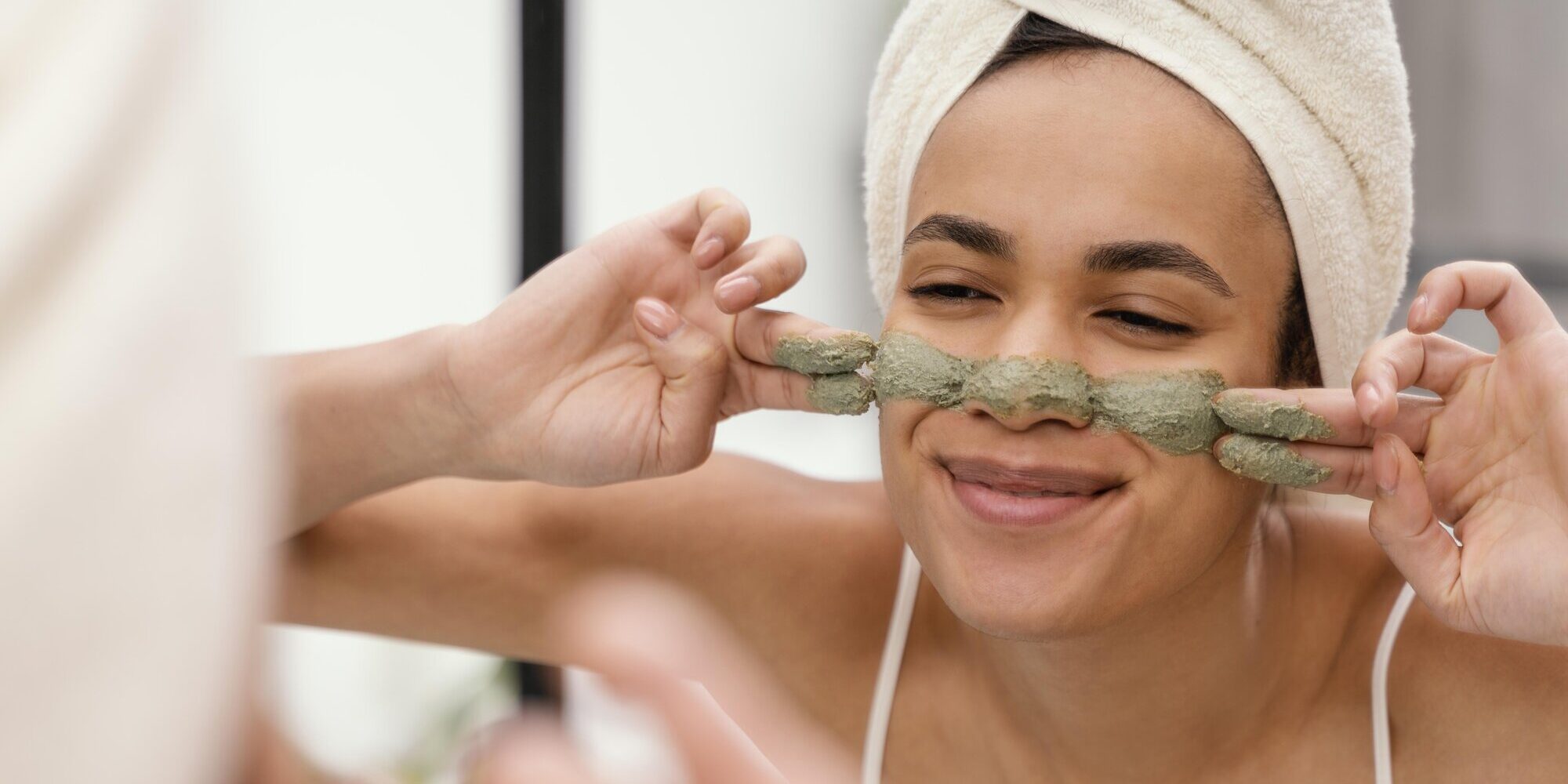 Frau mit Handtuch um den Kopf zeigt zufrieden eine Gesichtsmaske aus grünem Material im Badezimmer.