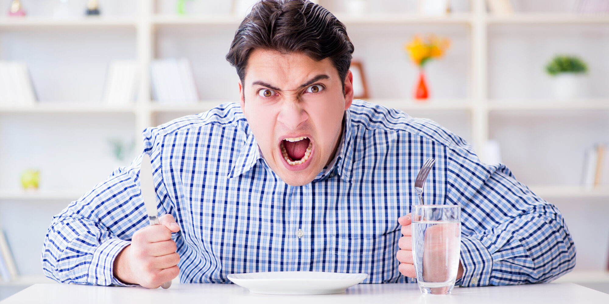 Ein wütender Mann hält ein Messer und eine Gabel über einem leeren Teller, während er an einem Tisch sitzt.