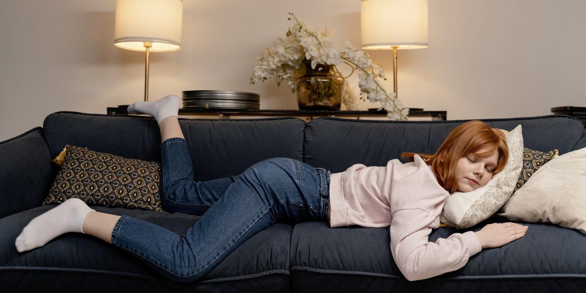 powernap.jpg Eine junge Frau mit roten Haaren liegt entspannt auf einem blauen Sofa, umgeben von Kissen und Lampen.