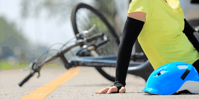 Eine Person in einem gelben Shirt sitzt auf der Straße neben einem blauen Fahrradhelm und einem umgekippten Fahrrad.