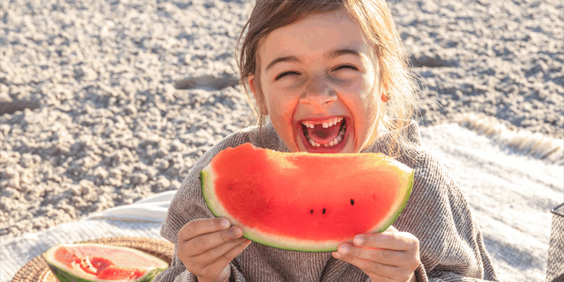 Obst_psychische-Gesundheit.png Ein lachendes Kind hält ein großes Stück Wassermelone in der Hand, umgeben von einer sommerlichen Picknick-Atmosphäre.