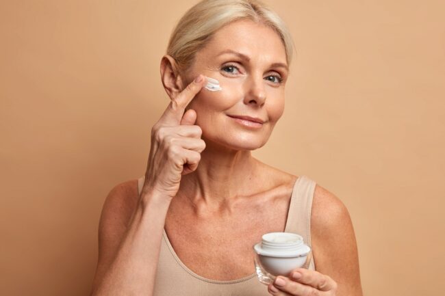Anti-Aging.jpg Eine Frau mit blondem Haar trägt Gesichtspflegecreme auf ihre Wange auf, während sie einen Cremetiegel in der Hand hält.