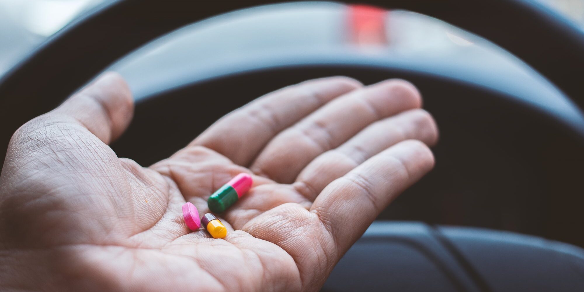 Arzneimittel_im_Strassenverkehr.jpg Eine Hand hält bunte Kapseln über dem Lenkrad eines Fahrzeugs.