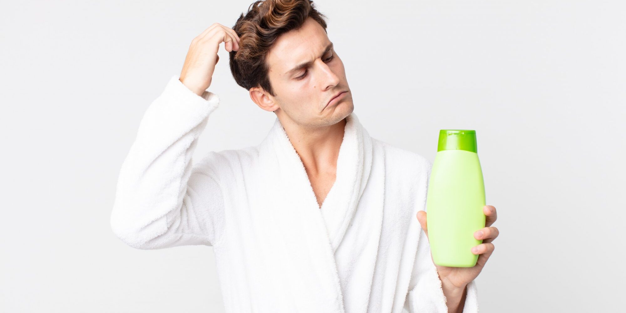 Trockene_Kopfhaut_Pflege.jpg Ein Mann in einem weißen Bademantel hält eine grüne Shampoo-Flasche und sieht nachdenklich aus.
