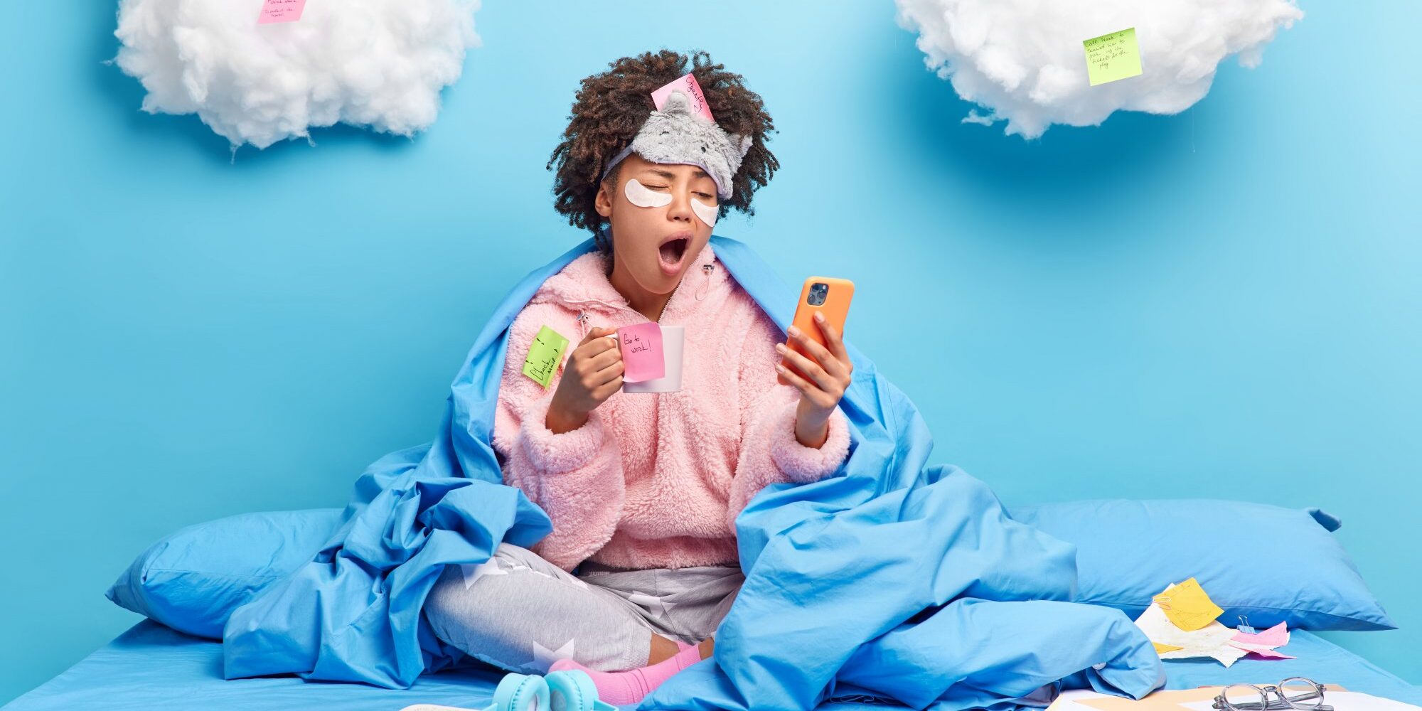 Soforttipps_gegen_Muedigkeit.jpg Eine Person in einem blauen Bett, umhüllt von einer Decke, schaut müde auf ihr Handy und hält eine Tasse in der Hand. Wolken mit Haftnotizen im Hintergrund.