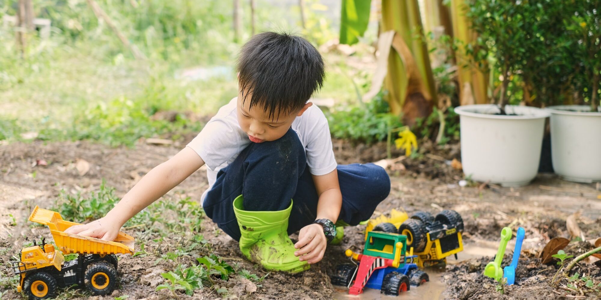 wie-viel-dreck-ist-gesund.jpg Ein Junge im Garten spielt mit verschiedenen Spielzeugfahrzeugen, darunter ein Lkw und ein Bagger, während er in matschigem Boden sitzt.