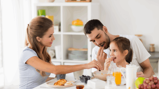 Familie beim gemeinsamen Frühstück an einem Tisch mit Obst, Pfannkuchen und Getränken.
