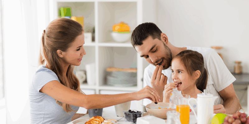 Familie beim gemeinsamen Frühstück an einem Tisch mit Obst, Pfannkuchen und Getränken.
