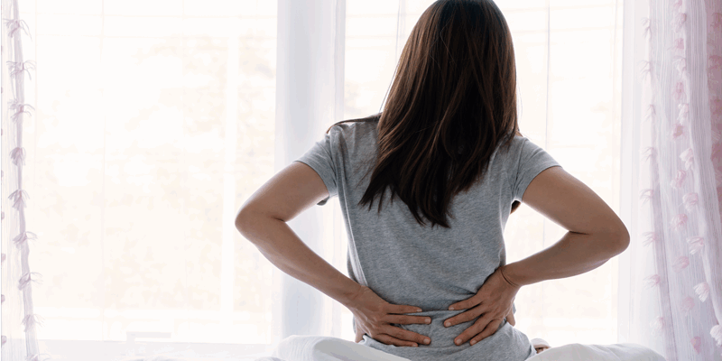 Eine Person mit langen Haaren sitzt auf einem Bett und hält sich den Rücken vor einem Fenster.