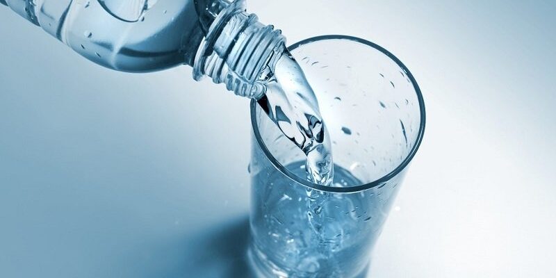 Wasserglas_WEB.jpg Eine Wasserflasche schüttet frisches Wasser in ein Glas, das auf einer hellen, blauen Oberfläche steht.