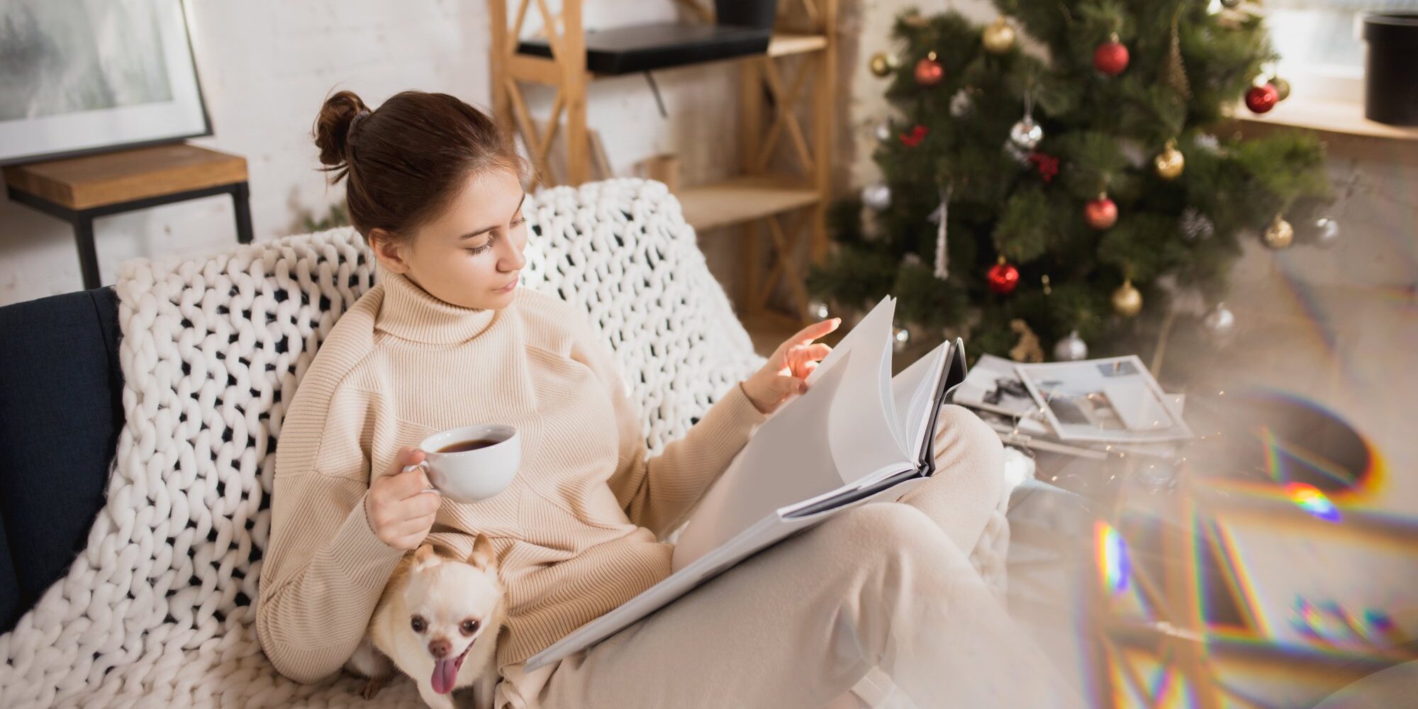 Eine Frau in bequemer Kleidung sitzt auf einem Sofa, liest in einem Buch und hält eine Tasse Kaffee, während ein Hund bei ihr sitzt.