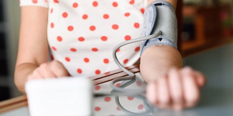 Eine Person mit einem Blutdruckmessgerät am Arm, das auf einem Tisch liegt. Im Hintergrund ein unscharfer Raum.
