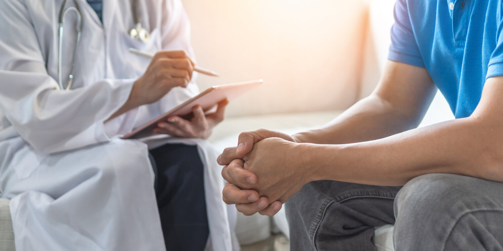 Arzt in weißem Kittel notiert auf einem Tablet, während ein Patient mit zusammengefalteten Händen sitzt.