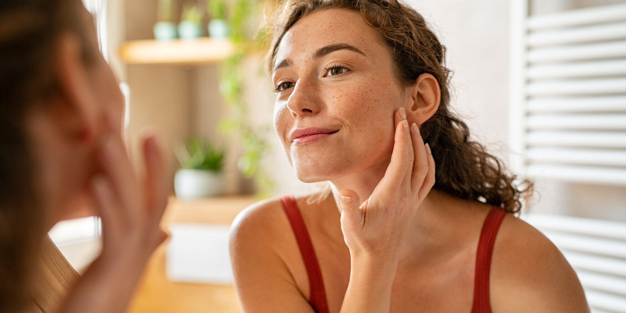 gesunde-haut.jpeg Frau mit lockigem Haar, die im Spiegel ihre Haut berührt, sanfte Beleuchtung und Pflanzen im Hintergrund.