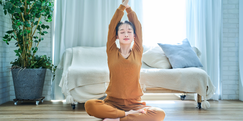Vagusnerv.png Eine Person in orangefarbenem Yoga-Outfit sitzt im Schneidersitz und streckt die Arme nach oben, umgeben von heller, luftiger Wohnungseinrichtung.