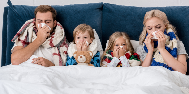 Eine Familie mit zwei Kindern kuschelt sich in Decken auf einer Couch, alle halten Taschentücher vor den Nasen.