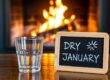 Ein Glas Wasser steht neben einer Tafel mit der Aufschrift 'DRY JANUARY' vor einem Kaminfeuer.