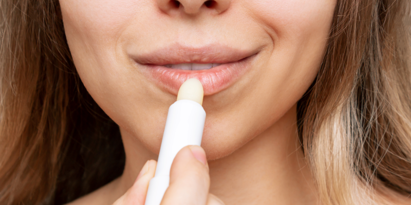 Lippenpflege.png Frau trägt einen Lippenbalsam auf ihre Lippen auf, der in einem weißen Stick präsentiert wird.