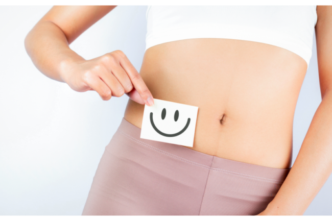 Eine Person hält eine Notiz mit einem lächelnden Gesicht vor den Bauch, auf weißem Hintergrund.