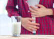 Ein Glas Milch steht auf einem Tisch, während eine Person im Hintergrund den Bauch hält.