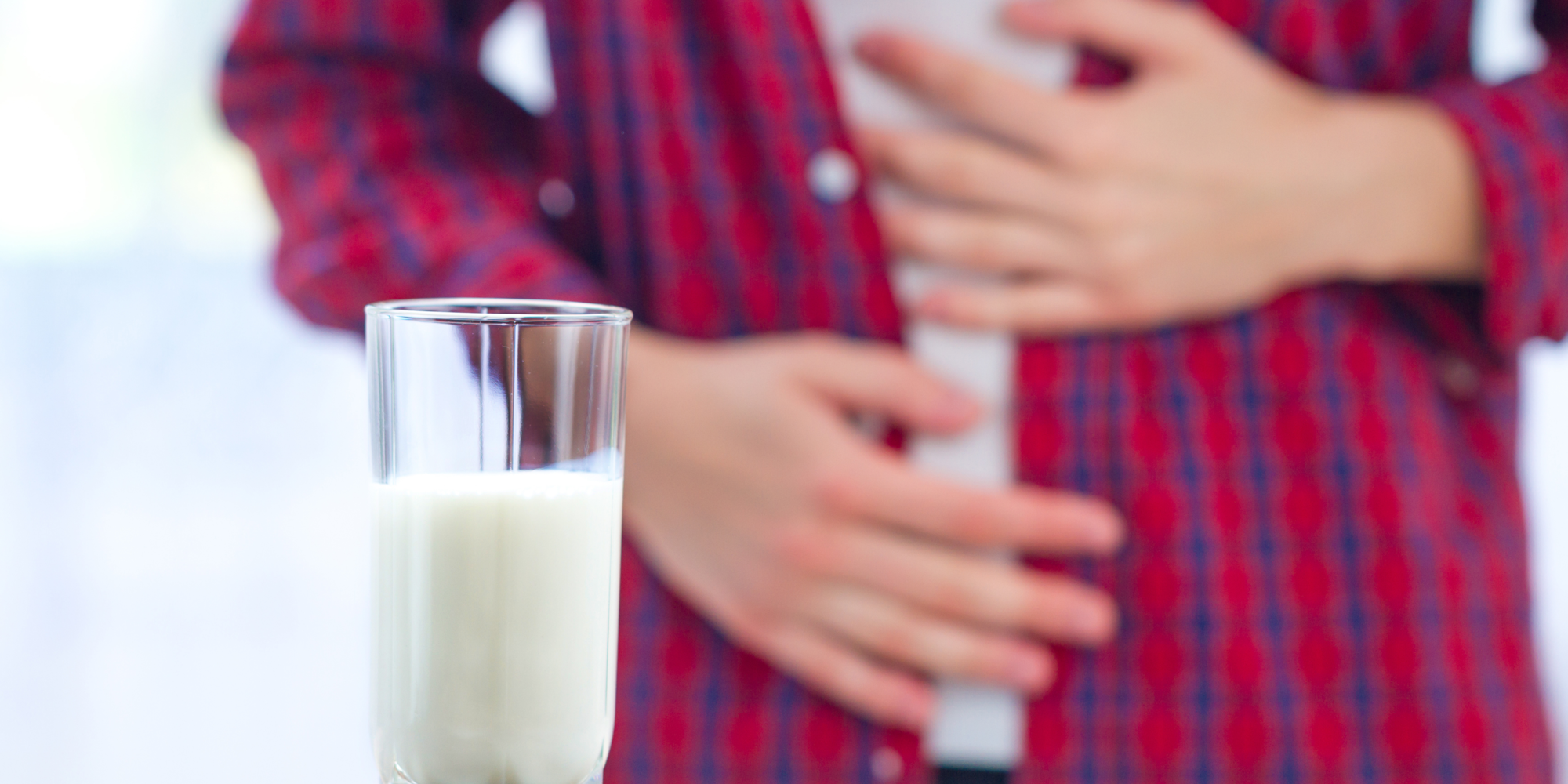 Ein Glas Milch steht auf einem Tisch, während eine Person im Hintergrund den Bauch hält.
