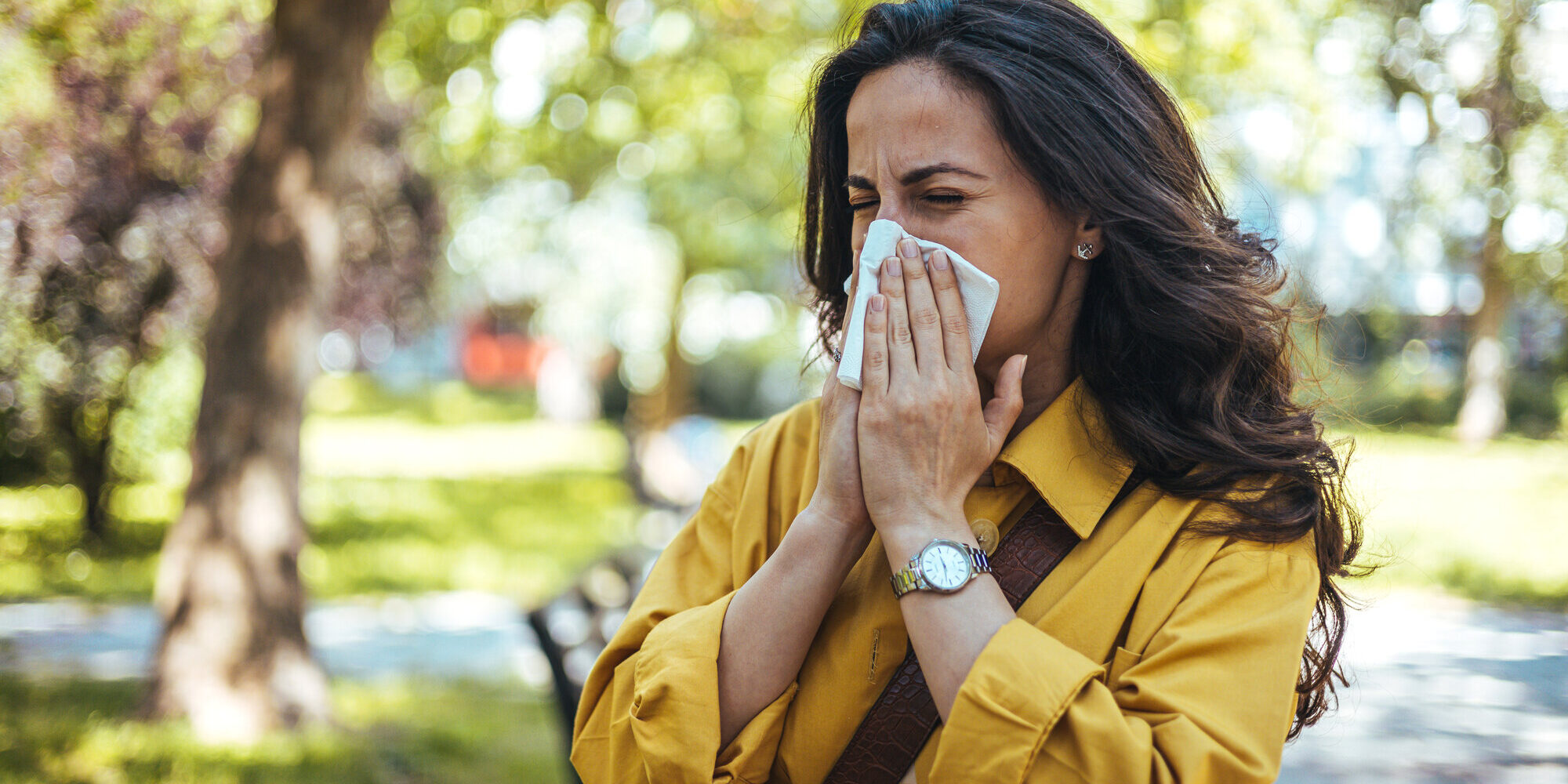 Frau in einem gelben Hemd, die sich mit einem Taschentuch die Nase putzt, umgeben von grüner Parklandschaft.
