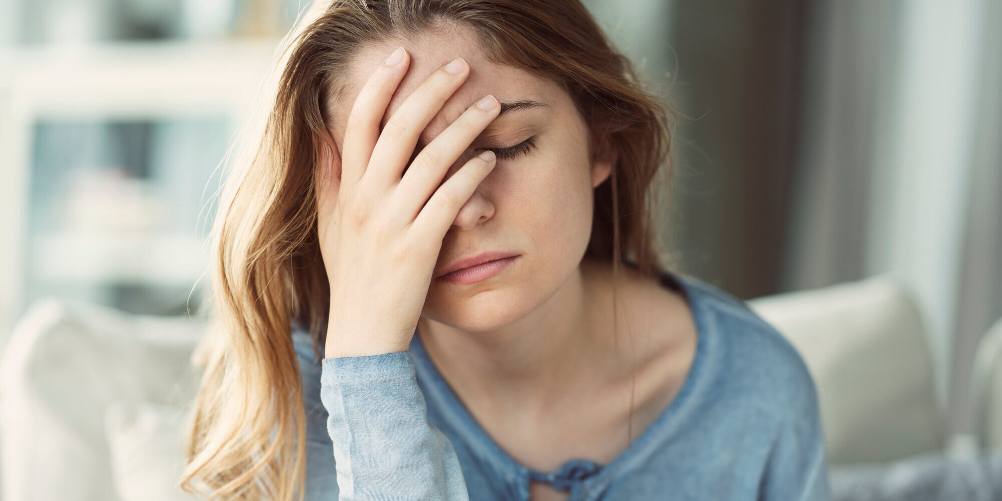 chronische-entzuendungen.jpeg Junge Frau mit langen Haaren und blauem Oberteil, die frustriert ihre Hand an die Stirn legt, im Innenraum sitzend.